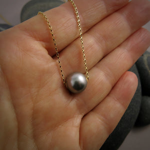 A pearl necklace is displayed on a hand.  The round, gray pearl is suspended on a yellow gold filled rolo chain that slides through the centre of the pearl.