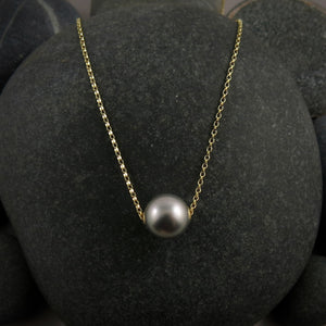 A pearl necklace is displayed on a black beach rock.  The round, gray pearl is suspended on a yellow gold filled rolo chain that slides through the centre of the pearl.
