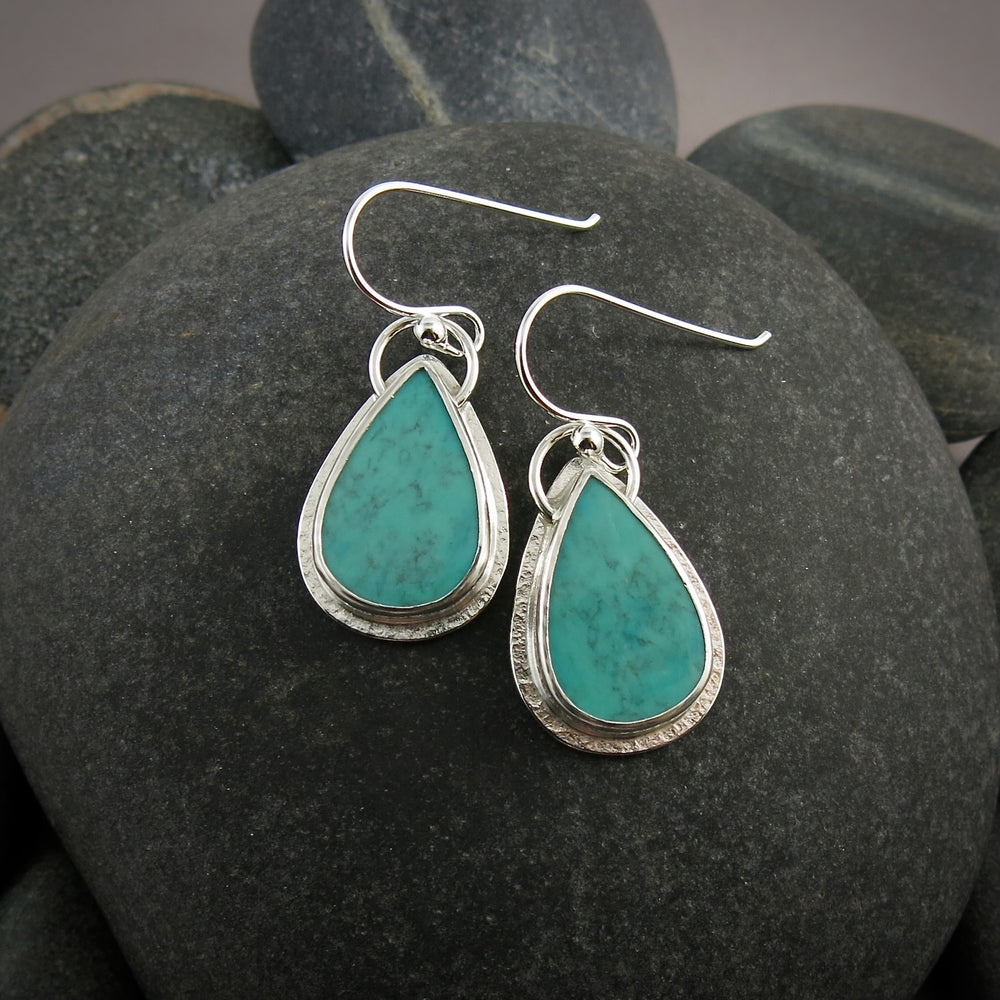A pair of teardrop shaped turquoise earrings are displayed on a black beach rock.  The turquoise are a lovely natural blue-green and feature a natural matrix pattern.  They are set in sterling silver.