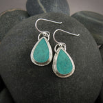 A pair of teardrop shaped turquoise earrings are displayed on a black beach rock.  The turquoise are a lovely natural blue-green and feature a natural matrix pattern.  They are set in sterling silver.
