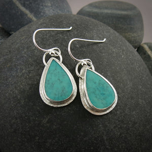 A pair of teardrop shaped turquoise earrings are displayed on a black beach rock.  The turquoise are a lovely natural blue-green and feature a natural matrix pattern.  They are set in sterling silver.