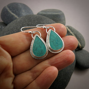 A pair of teardrop shaped turquoise earrings are displayed on a hand.  The turquoise are a lovely natural blue-green and feature a natural matrix pattern.  They are set in sterling silver.
