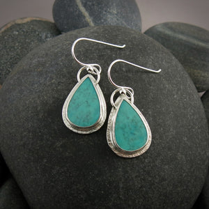 A pair of teardrop shaped turquoise earrings are displayed on a black beach rock.  The turquoise are a lovely natural blue-green and feature a natural matrix pattern.  They are set in sterling silver.