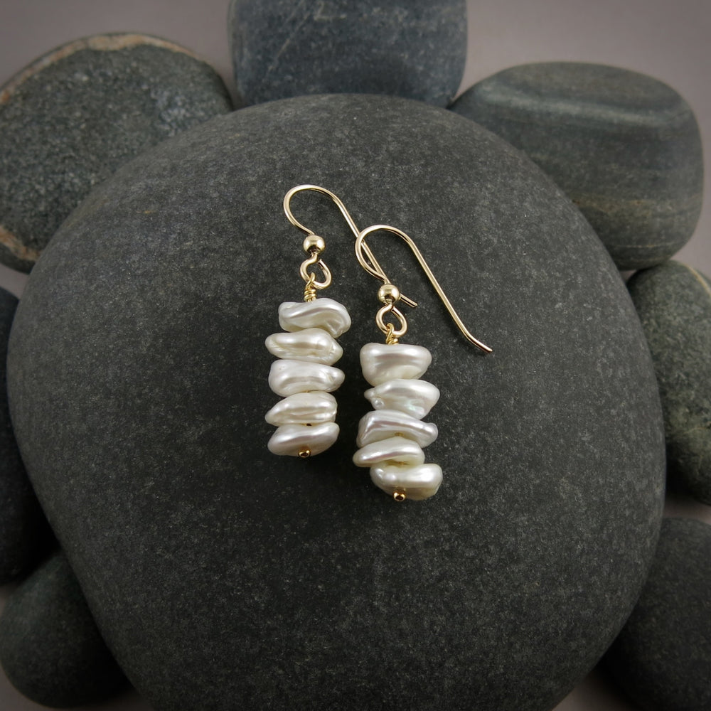 A pair of pearl earrings are displayed on a black beach rock.  The earrings feature a stack of five organically shaped, white keshi pearls on gold ear wires.