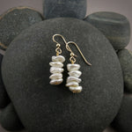 A pair of pearl earrings are displayed on a black beach rock.  The earrings feature a stack of five organically shaped, white keshi pearls on gold ear wires.
