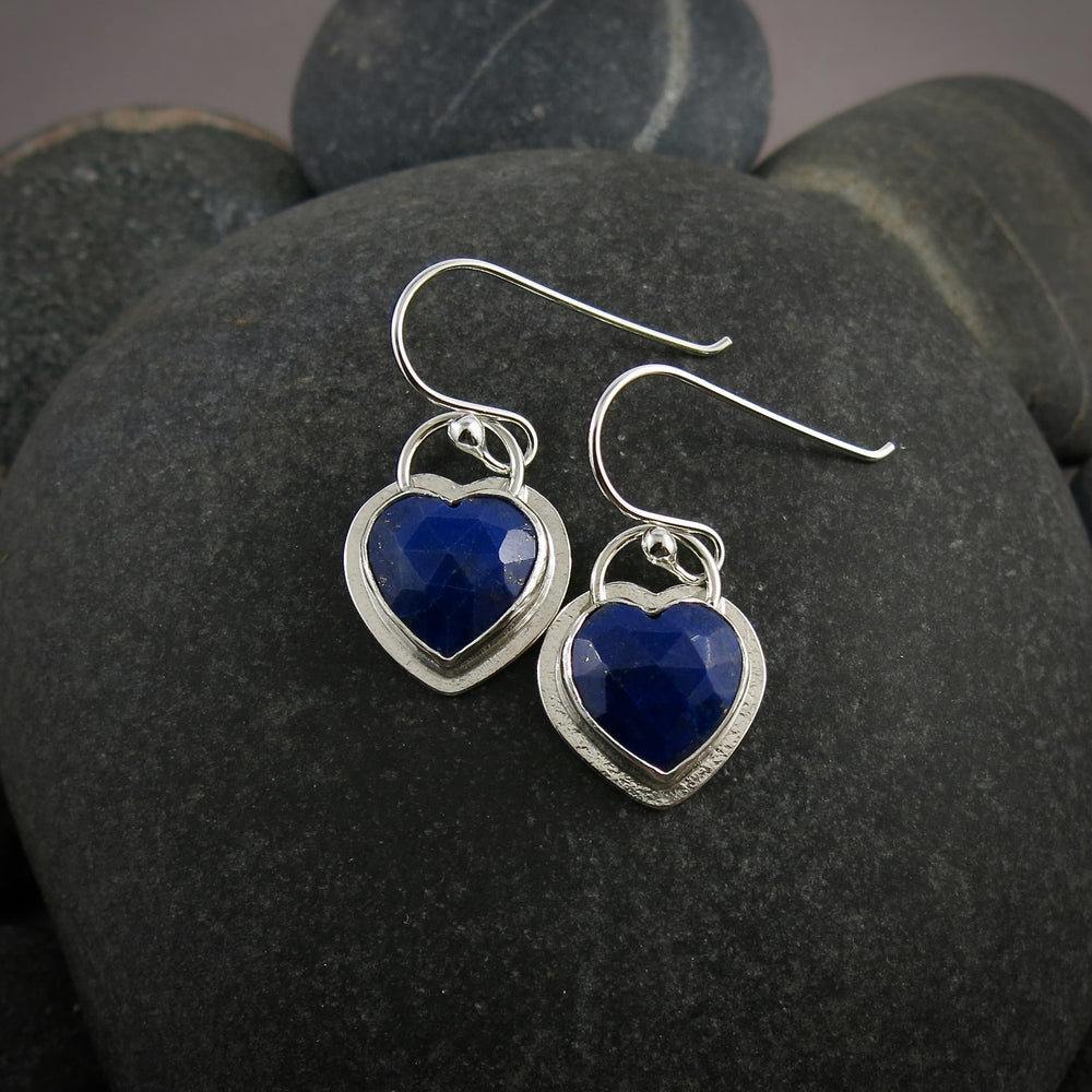 A pair of heart shaped royal blue lapis lazuli earrings are displayed on a black beach rock.  The gemstones are set in a sterling silver setting with a subtly textured "halo" of silver surrounding the gems.