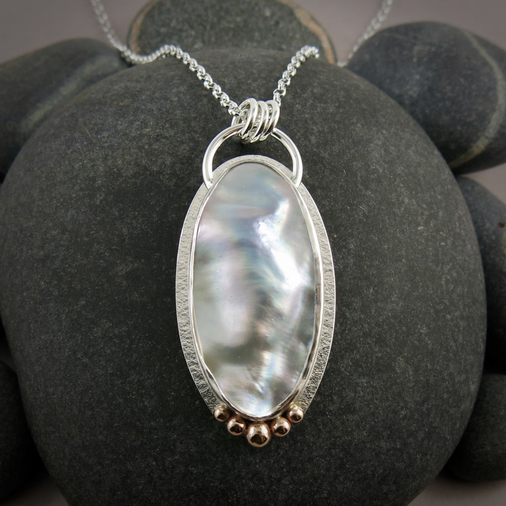 A large mabe pearl statement necklace is displayed against a black beach rock.  The necklace features an oval pearl set in sterling silver with give 14K gold ball accents at the bottom of the pendant.