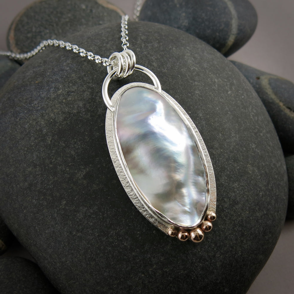 A large mabe pearl statement necklace is displayed against a black beach rock.  The necklace features an oval pearl set in sterling silver with give 14K gold ball accents at the bottom of the pendant.