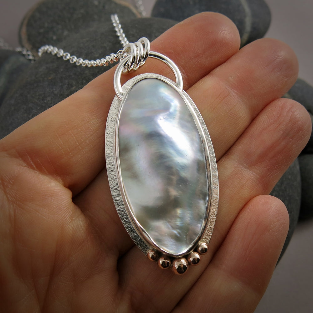 A large mabe pearl statement necklace is displayed on a hand.  The necklace features an oval pearl set in sterling silver with give 14K gold ball accents at the bottom of the pendant.