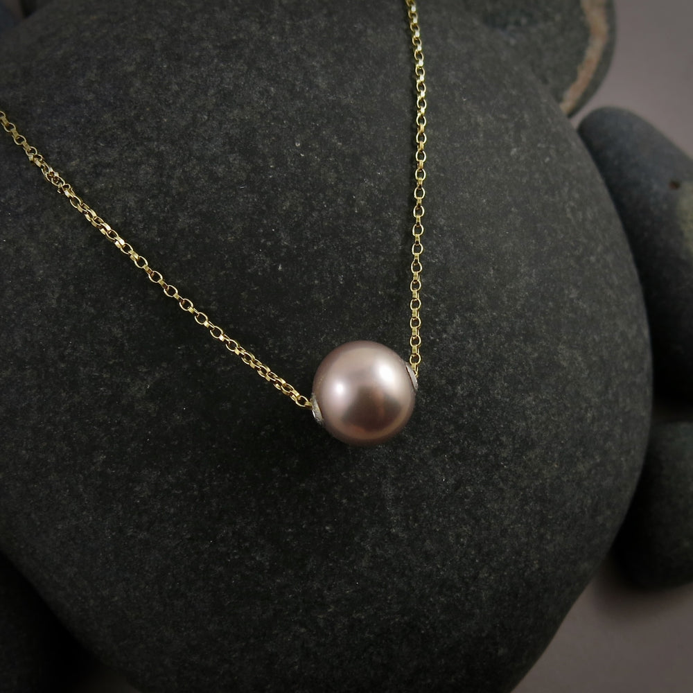 A pearl necklace is displayed on a black beach rock.  The round, lilac-pink pearl is suspended on a yellow gold filled rolo chain that slides through the centre of the pearl.