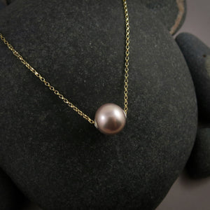 A pearl necklace is displayed on a black beach rock.  The round, lilac-pink pearl is suspended on a yellow gold filled rolo chain that slides through the centre of the pearl.