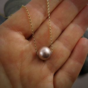 A pearl necklace is displayed on a hand.  The round, lilac-pink pearl is suspended on a yellow gold filled rolo chain that slides through the centre of the pearl.