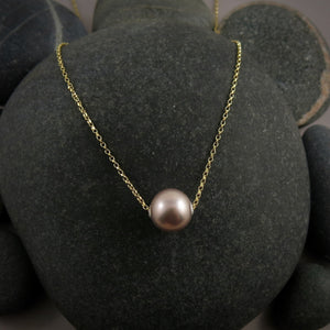 A pearl necklace is displayed on a black beach rock.  The round, lilac-pink pearl is suspended on a yellow gold filled rolo chain that slides through the centre of the pearl.