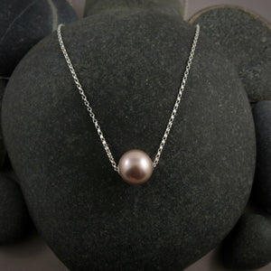 A pearl necklace is displayed on a black beach rock.  The round, lilac-pink pearl is suspended on a sterling silver rolo chain that slides through the centre of the pearl.