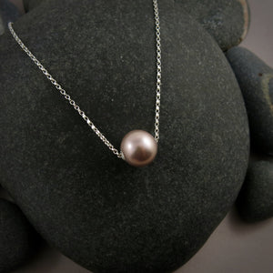A pearl necklace is displayed on a black beach rock.  The round, lilac-pink pearl is suspended on a sterling silver rolo chain that slides through the centre of the pearl.