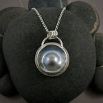 A stunningly lustrous mabe pearl necklace is displayed on a black beach rock.  The mabe pearl, also known as a blister pearl, displays a rainbow of colours on a mostly silver surface.  The pearl is set in a handcrafted sterling silver setting with a bezel that follows the organic shape of the pearl.  