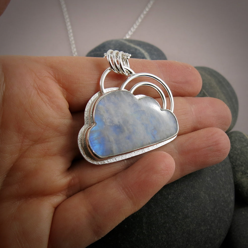 A handcrafted sterling silver necklace by Mikel Grant Jewellery.  A rainbow moonstone shaped like a cloud is set in sterling silver with a "silver lining" of silver surrounding the gem.  A double "rainbow" of curved silver wire rests atop the cloud.