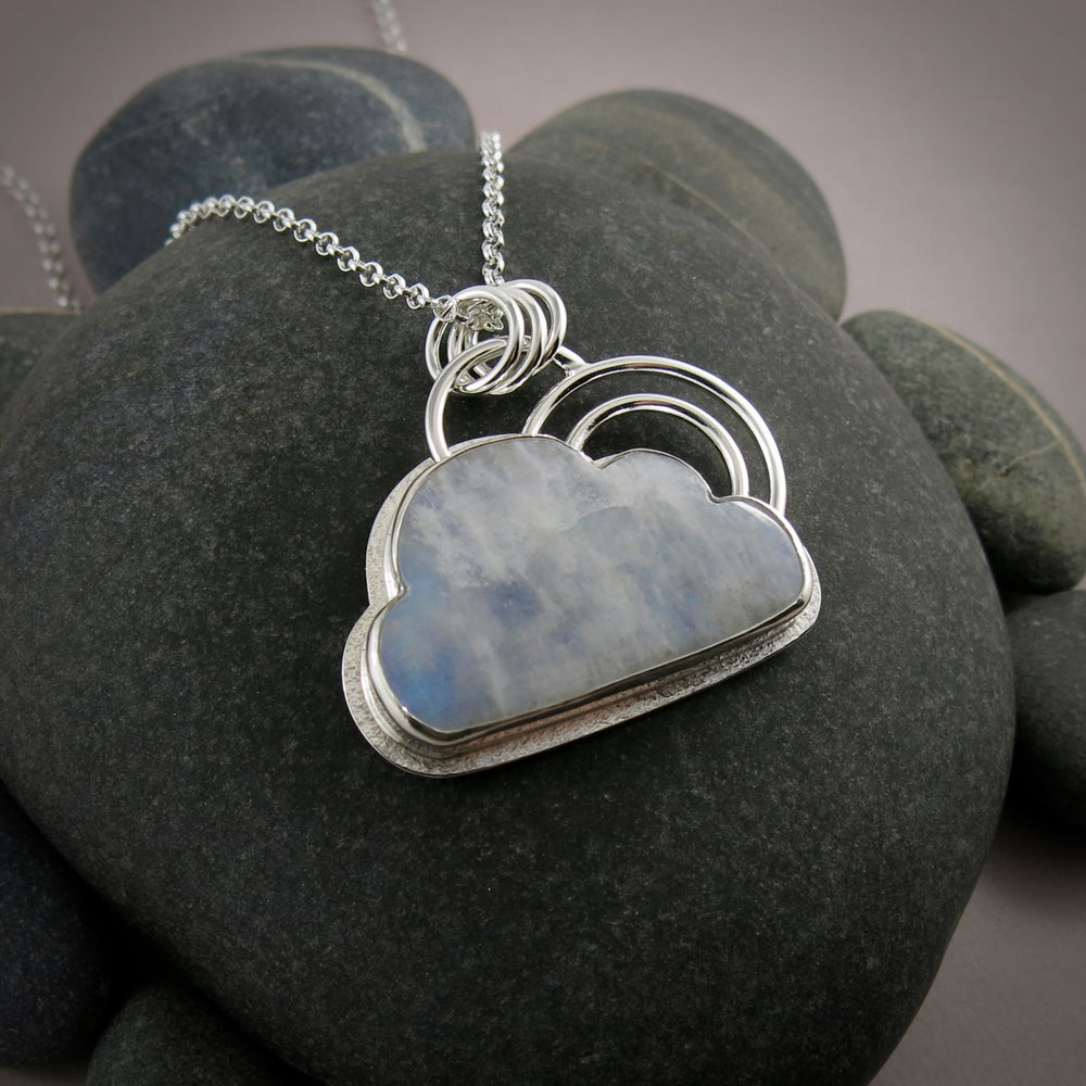 A handcrafted sterling silver necklace by Mikel Grant Jewellery.  A rainbow moonstone shaped like a cloud is set in sterling silver with a "silver lining" of silver surrounding the gem.  A double "rainbow" of curved silver wire rests atop the cloud.