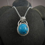 A bright blue turquoise pendant is displayed on a black beach rock.  The pendant features an oval shaped Morenci turquoise in a bright blue colour with specks of silver pyrite.  The gem is set in a sterling silver setting with a "halo" of textured silver around the bezel set gem and a double silver "rainbow" of silver wire above.   Three silver rings attach the chain to the rainbow. 