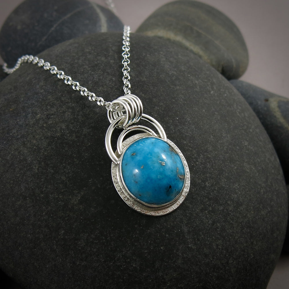 A bright blue turquoise pendant is displayed on a black beach rock.  The pendant features an oval shaped Morenci turquoise in a bright blue colour with specks of silver pyrite.  The gem is set in a sterling silver setting with a "halo" of textured silver around the bezel set gem and a double silver "rainbow" of silver wire above.   Three silver rings attach the chain to the rainbow. 