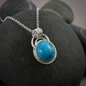 A bright blue turquoise pendant is displayed on a black beach rock.  The pendant features an oval shaped Morenci turquoise in a bright blue colour with specks of silver pyrite.  The gem is set in a sterling silver setting with a "halo" of textured silver around the bezel set gem and a double silver "rainbow" of silver wire above.   Three silver rings attach the chain to the rainbow. 