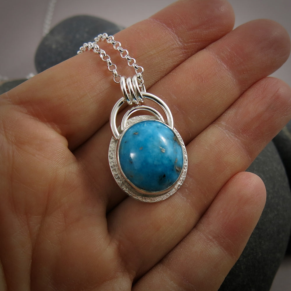 A bright blue turquoise pendant is displayed on a hand.  The pendant features an oval shaped Morenci turquoise in a bright blue colour with specks of silver pyrite.  The gem is set in a sterling silver setting with a "halo" of textured silver around the bezel set gem and a double silver "rainbow" of silver wire above.   Three silver rings attach the chain to the rainbow. 