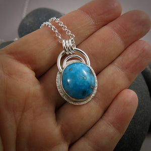 A bright blue turquoise pendant is displayed on a hand.  The pendant features an oval shaped Morenci turquoise in a bright blue colour with specks of silver pyrite.  The gem is set in a sterling silver setting with a "halo" of textured silver around the bezel set gem and a double silver "rainbow" of silver wire above.   Three silver rings attach the chain to the rainbow. 