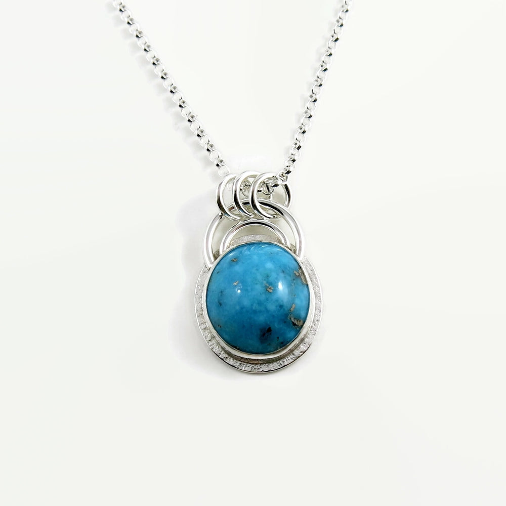 A bright blue turquoise pendant is displayed on a white background.  The pendant features an oval shaped Morenci turquoise in a bright blue colour with specks of silver pyrite.  The gem is set in a sterling silver setting with a "halo" of textured silver around the bezel set gem and a double silver "rainbow" of silver wire above.   Three silver rings attach the chain to the rainbow. 