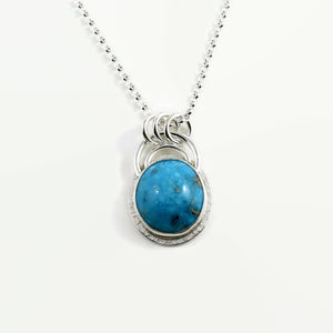 A bright blue turquoise pendant is displayed on a white background.  The pendant features an oval shaped Morenci turquoise in a bright blue colour with specks of silver pyrite.  The gem is set in a sterling silver setting with a "halo" of textured silver around the bezel set gem and a double silver "rainbow" of silver wire above.   Three silver rings attach the chain to the rainbow. 