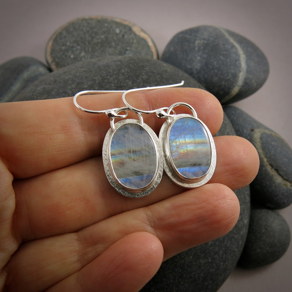 A pair of oval rainbow moonstone earrings are displayed on a hand.  The colourful gemstones are set in sterling silver.