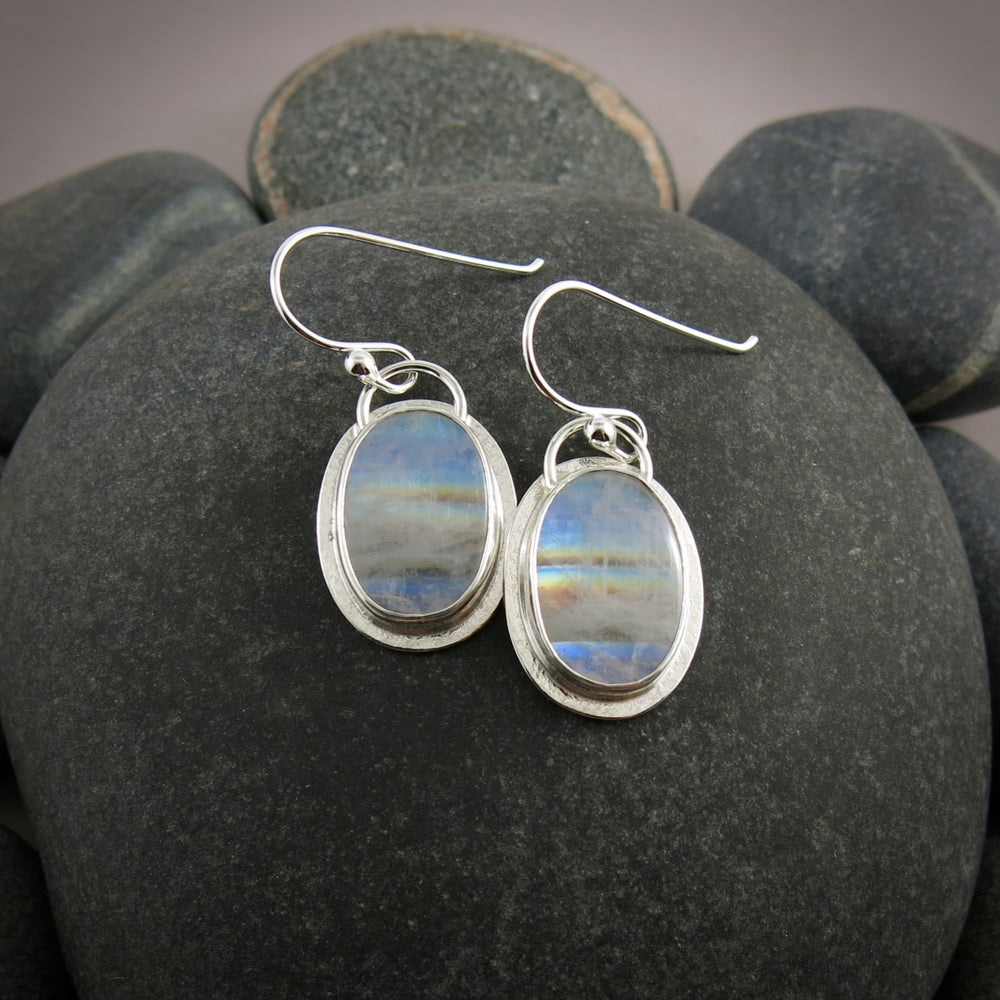 A pair of oval rainbow moonstone earrings are displayed on a black beach rock.  The colourful gemstones are set in sterling silver.