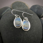 A pair of oval rainbow moonstone earrings are displayed on a black beach rock.  The colourful gemstones are set in sterling silver.