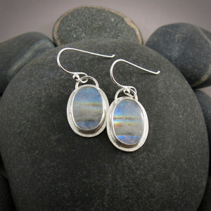 A pair of oval rainbow moonstone earrings are displayed on a black beach rock.  The colourful gemstones are set in sterling silver.