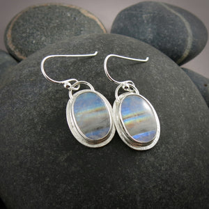 A pair of oval rainbow moonstone earrings are displayed on a black beach rock.  The colourful gemstones are set in sterling silver.