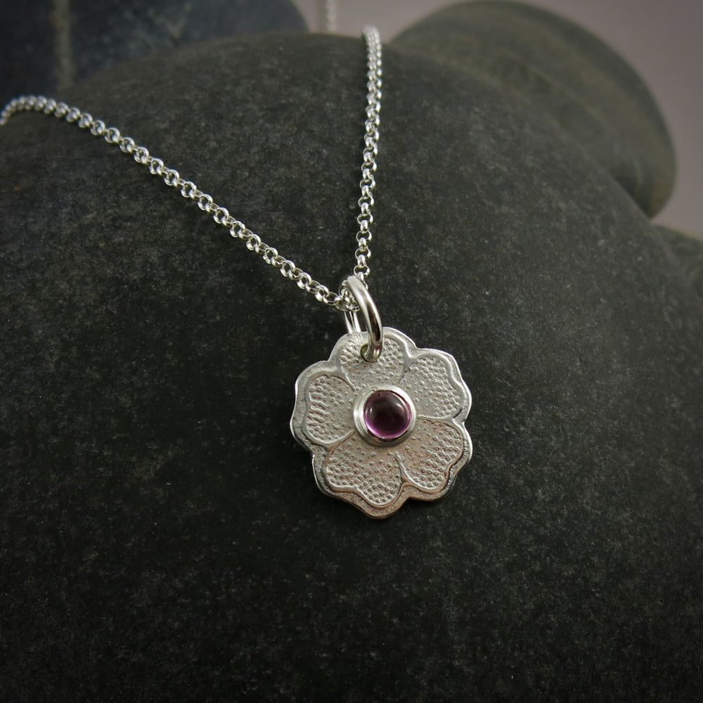 Silver necklace with a floral pendant featuring a pink gemstone on a dark surface.