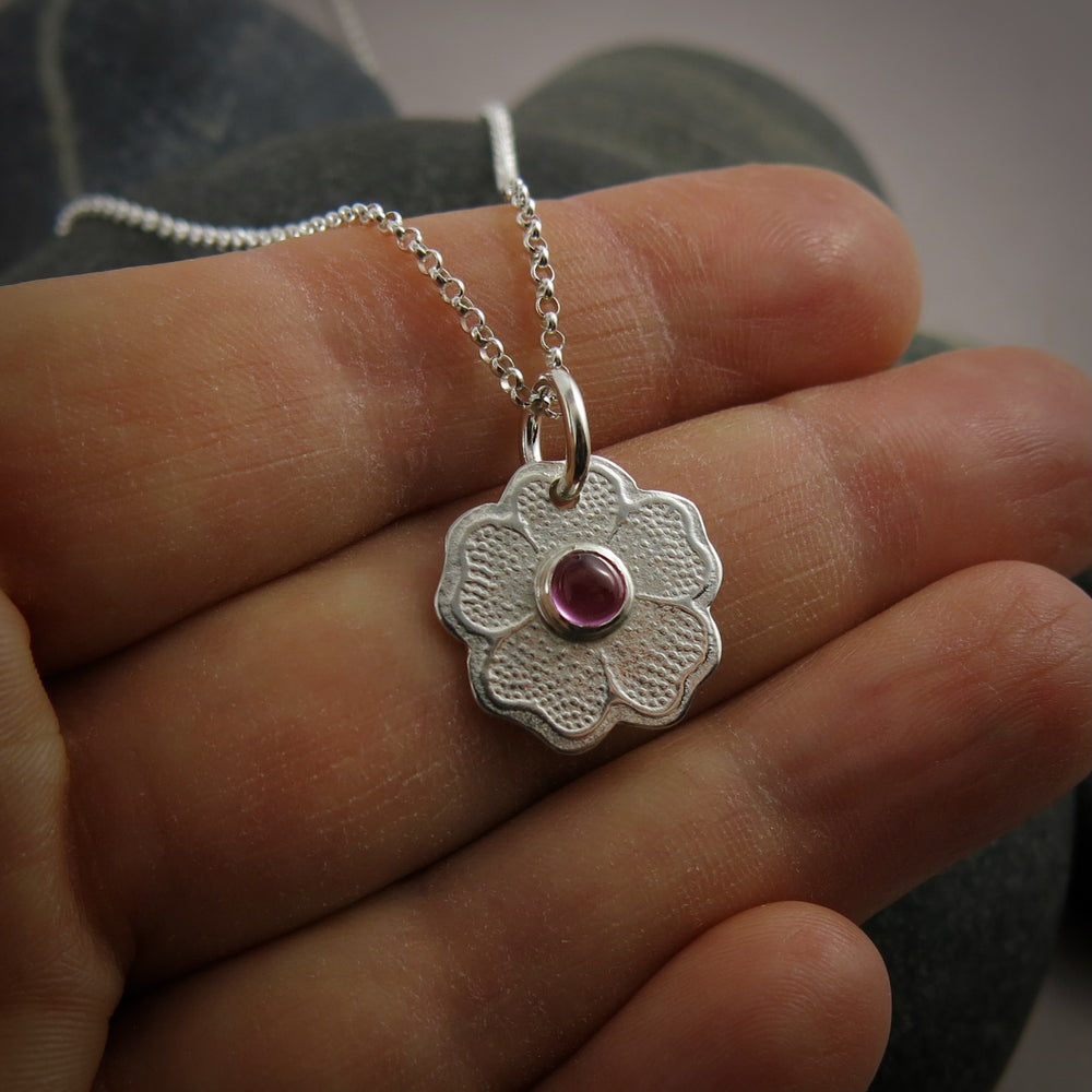 Silver necklace with a floral pendant featuring a pink gemstone held between fingers.