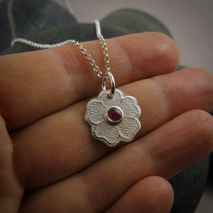 Silver necklace with a floral pendant featuring a pink gemstone held between fingers.