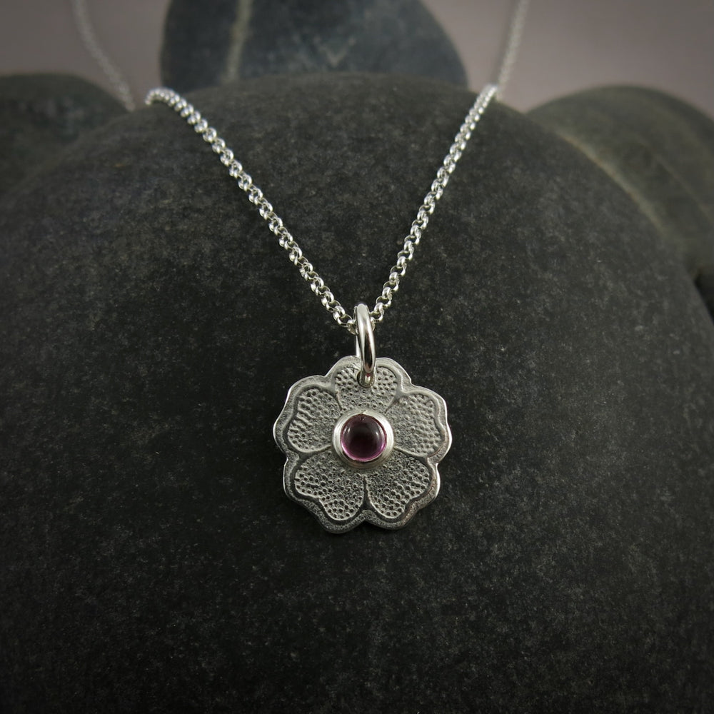 Silver necklace with a floral pendant featuring a pink gemstone on a dark gray surface.