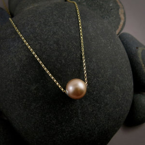 A pearl necklace is displayed on a black beach rock.  The round, peachy-pink pearl is suspended on a 14K gold filled rolo chain that slides through the centre of the pearl.