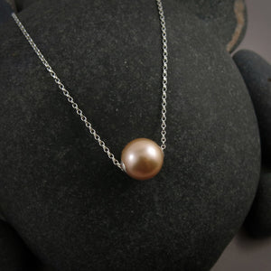 A pearl necklace is displayed on a black beach rock.  The round, peachy-pink pearl is suspended on a sterling silver rolo chain that slides through the centre of the pearl.