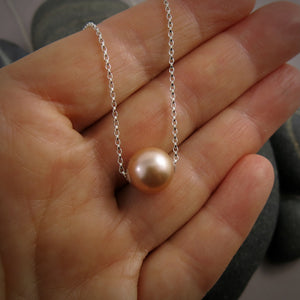 A pearl necklace is displayed on a hand.  The round, peachy-pink pearl is suspended on a sterling silver rolo chain that slides through the centre of the pearl.
