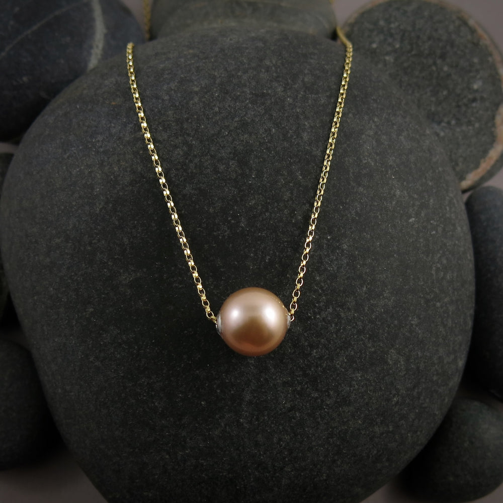 A pearl necklace is displayed on a black beach rock.  The round, peachy-pink pearl is suspended on a yellow gold filled rolo chain that slides through the centre of the pearl.