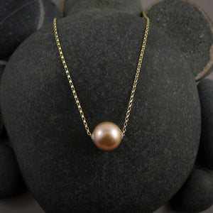 A pearl necklace is displayed on a black beach rock.  The round, peachy-pink pearl is suspended on a yellow gold filled rolo chain that slides through the centre of the pearl.