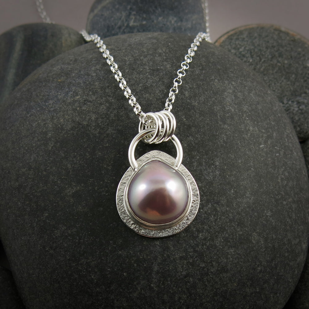 A pink teardrop shaped mabe pearl necklace in a silver setting is displayed against a black beach rock.
