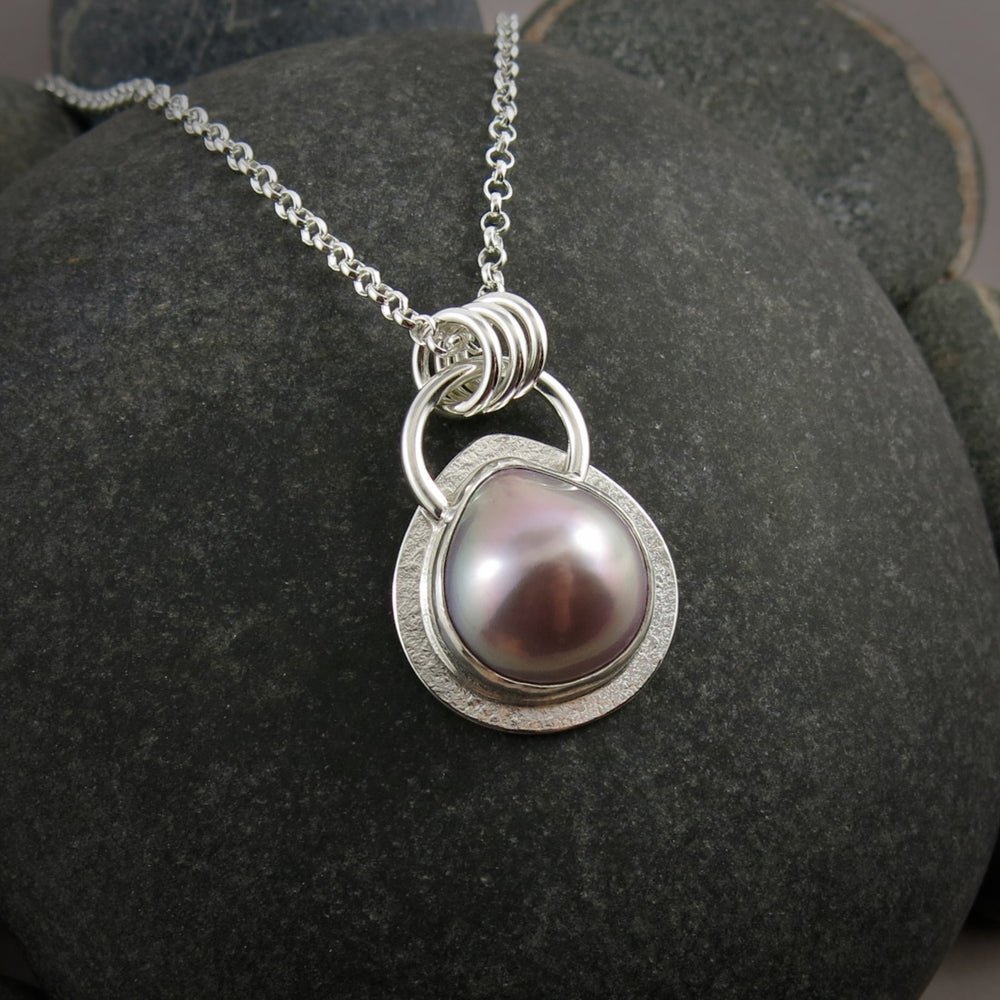 A pink teardrop shaped mabe pearl necklace in a silver setting is displayed against a black beach rock.
