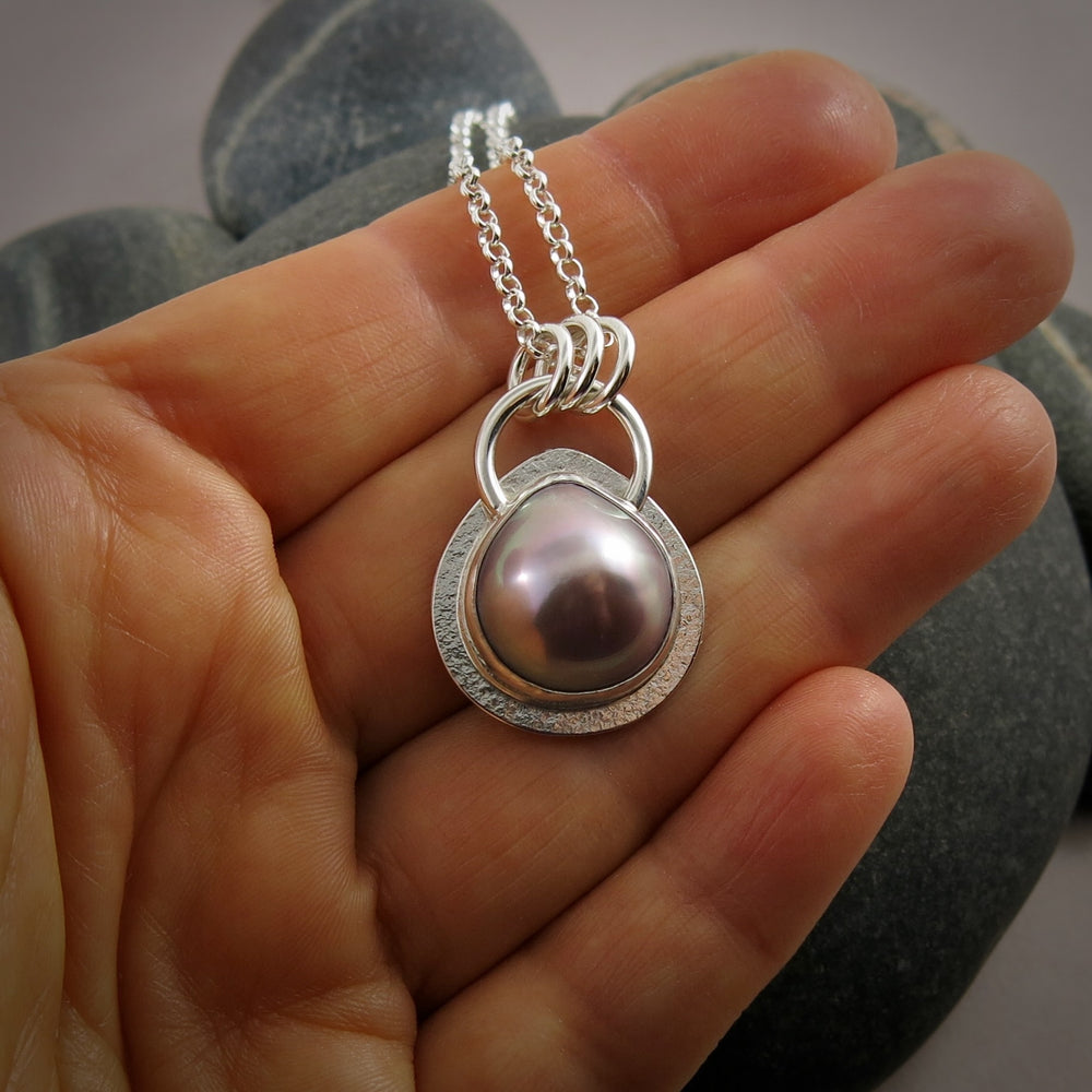 A pink teardrop shaped mabe pearl necklace in a silver setting is displayed on a hand.