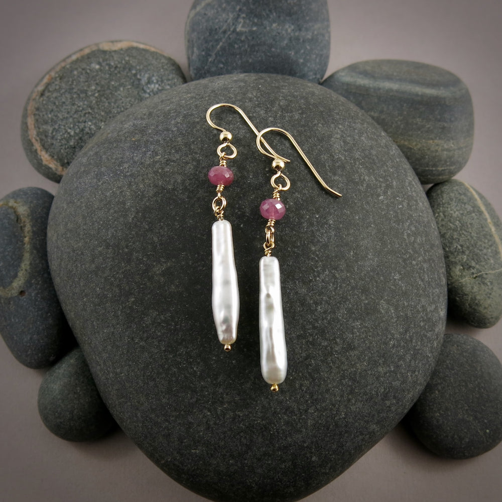 A pair of gemstone & pearl earrings are displayed on a black beach rock.  The earrings feature a faceted pink sapphire bead atop lustrous white stick pearls.  The ear wires and headpins are 14K gold filled.