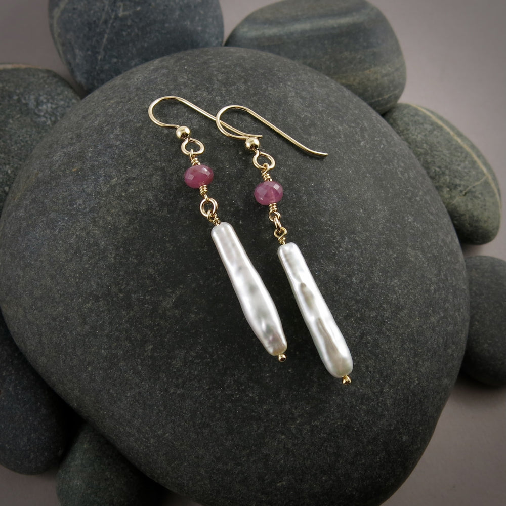 A pair of gemstone & pearl earrings are displayed on a black beach rock.  The earrings feature a faceted pink sapphire bead atop lustrous white stick pearls.  The ear wires and headpins are 14K gold filled.