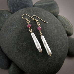 A pair of gemstone & pearl earrings are displayed on a black beach rock.  The earrings feature a faceted pink sapphire bead atop lustrous white stick pearls.  The ear wires and headpins are 14K gold filled.