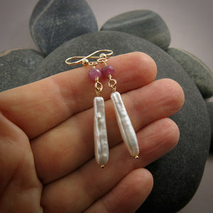 A pair of gemstone & pearl earrings are displayed on a hand.  The earrings feature a faceted pink sapphire bead atop lustrous white stick pearls.  The ear wires and headpins are 14K gold filled.